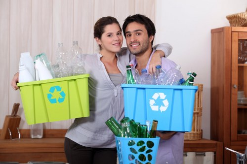 A young couple standing indoors in a kitchen or living space, each holding a plastic recycling bin filled with empty glass bottles; the woman’s green bin contains clear glass bottles, while the man’s blue bin, which features a white recycling symbol, holds additional glass bottles. They are smiling and appear to be in the process of sorting recyclable waste for disposal or collection, with the woman’s arm around the man’s shoulder. The background includes a light-colored wooden or laminate wall, a wooden sideboard, and a few decorative items such as a glass container or vase, with natural or ambient lighting illuminating the scene. This image visually represents environmentally conscious rubbish sorting, relevant to waste management and recycling services in areas like Honor Oak or the surrounding London boroughs, associated with professional rubbish removal efforts by Garden Clearance Honor Oak.