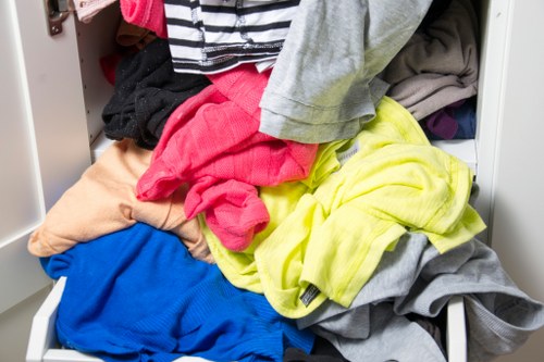 A partially open white wardrobe drawer filled with a variety of crumpled and piled clothes, including brightly colored items such as a pink garment, a neon yellow piece, and blue and grey fabrics, with some darker clothing visible underneath. The setting appears to be in a domestic environment, with some clothing spilling out of the drawer and a glimpse of the wardrobe's interior. The scene highlights clutter typical of laundry or clothing disposal, relevant to rubbish removal services such as those offered by Garden Clearance Honor Oak, supporting local waste management for homes in the Honor Oak area near London.