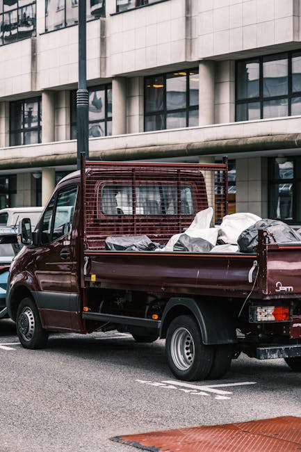 A small burgundy flatbed truck parked on an urban street in front of a modern office building with large glass windows and a concrete facade. The truck's cargo area is loaded with a variety of rubbish